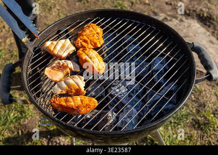 Filets de poulet grillant sur barbecue au charbon de bois. Gros plan de filets de poulet marinés cuisant sur un gril au charbon de bois à l'extérieur, avec des pinces en métal, des charbons chauds Banque D'Images