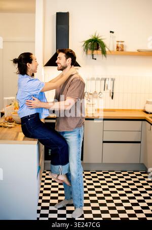 Un couple profite d’un moment ludique dans leur cuisine. La femme est assise sur le comptoir tandis que l'homme se tient proche, à la fois souriant et montrant de l'affection dans un b. Banque D'Images