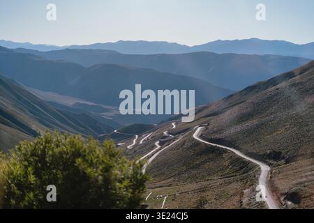 La route sinueuse traverse un terrain accidenté de montagne. Les pics distants s'estompent dans un horizon bleu brumeux. La lumière du soleil baigne les pentes d'une lueur chaude et dorée. Vert luxuriant Banque D'Images