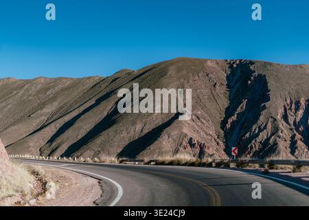 La route sinueuse traverse des montagnes escarpées. Le ciel bleu vif encadre le paysage sombre et ensoleillé. Les pentes sèches projettent de longues ombres nettes sur le terrain Banque D'Images