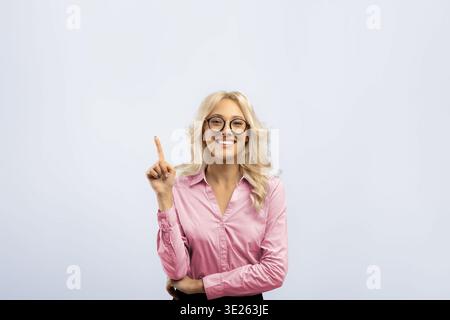 Femme sourit et pointe vers le haut tout en se tenant devant un fond Uni à l'intérieur Banque D'Images