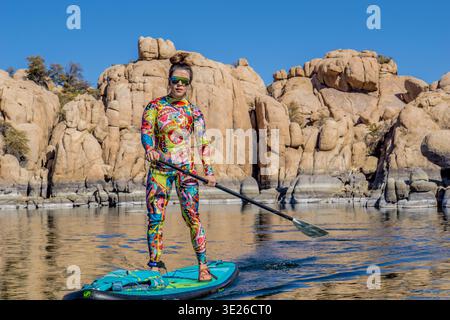 Une femme dans une tenue colorée se tient debout sur une planche de surf dans un plan d'eau. Elle tient une pagaie et elle s'amuse Banque D'Images