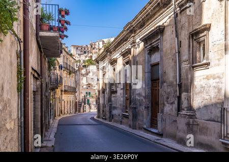 Vue sur une rue typique de Ragusa Ibla avec des bâtiments en calcaire, des balcons décorés de pots de fleurs et une architecture baroque. L'arrière-plan s'affiche Banque D'Images