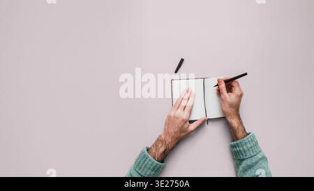 Mains masculines écrivant dans un petit cahier ouvert avec stylo noir. Fond lilas uniforme avec grand espace de copie sur la gauche. Deuxième stylo visible. Frais généraux v Banque D'Images