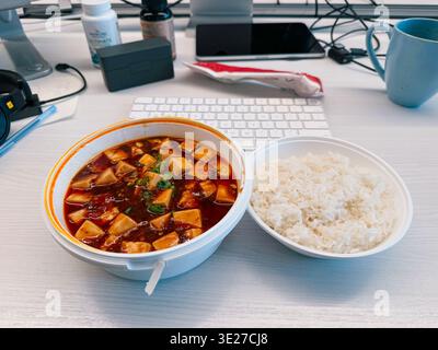 Bureau déjeuner avec tofu mapo et riz blanc à côté d'un clavier, téléphone et tasse à café dans un espace de travail décontracté Banque D'Images