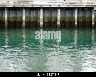 Les pieux de soutien métalliques et le mur de soutènement d'une promenade riveraine se reflètent dans les eaux vertes ondulantes du port de Vancouver Banque D'Images