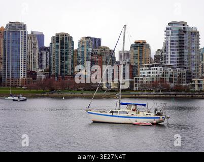 Un voilier blanc et bleu est amarré dans les eaux calmes de False Creek, contre la ligne d’horizon urbaine dense des gratte-ciel vitrés du centre-ville de Vancouver Banque D'Images
