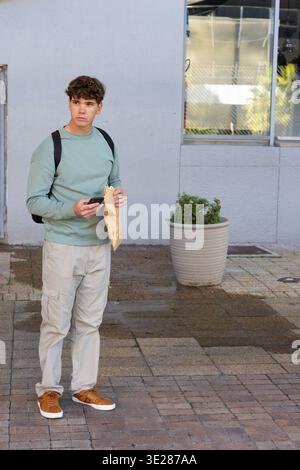 Adolescent homme debout sur la plaza tenant un smartphone et un sac en papier, portant un dessus de menthe et un sac à dos Banque D'Images