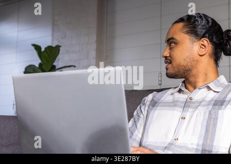 Homme asiatique assis sur un canapé gris dans le salon, tapant sur un ordinateur portable argenté, espace de copie Banque D'Images