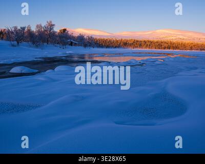 Une rivière coule devant les montagnes en Suède alors que la lumière du matin illumine le paysage. La neige recouvre le sol et l'eau reflète le soleil tôt Banque D'Images