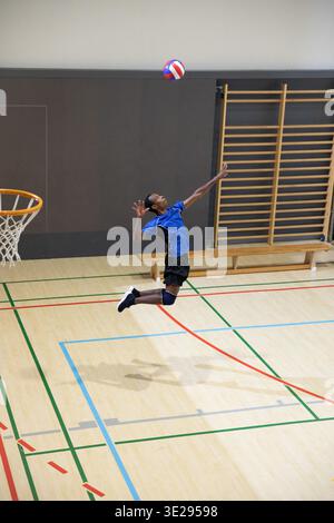 Homme adolescent afro-américain sautant frapper le volley rouge-blanc-bleu sur le terrain de gymnastique en maillot bleu. Athlétique, aéroporté, balle, cerceau, échelle, banc, lignes || modèle libéré Banque D'Images