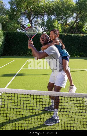 Papa afro-américain portant sa fille sur le dos et pratiquant le tennis avec raquette sur le court. Sports, famille, plein air, loisirs, dynamique, actif, connexion || modèle libéré Banque D'Images
