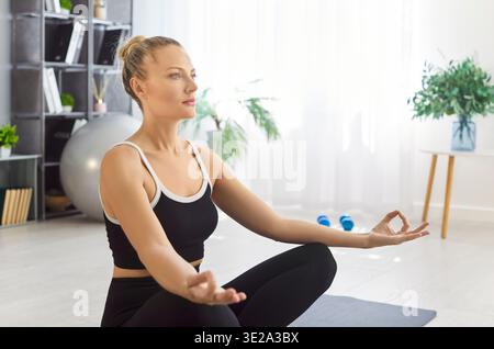 Yogi belle femme assise dans la pose de lotus sur tapis dans la salle de gym à la maison, méditant calmement formant mudra Banque D'Images