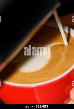Gros plan du lait versé dans un espresso fraîchement préparé, créant des motifs de crème onctueux dans une tasse rouge, préparation de café macro Banque D'Images