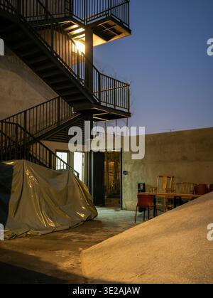 Une lumière chaleureuse rayonne d'une porte ouverte à côté d'un escalier extérieur en métal noir sur un bâtiment en béton sous un ciel crépusculaire bleu foncé. Evok Banque D'Images
