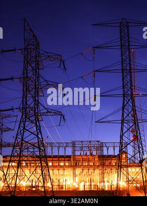 Vue nocturne d'une sous-station électrique avec des structures éclairées, des tours de transmission et des câbles sous un ciel bleu foncé pendant une longue exposition. Banque D'Images