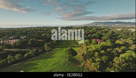 Portugal, Lisbonne : vue aérienne capturant un terrain de golf à Cascais, avec une verdure vibrante, des maisons aux toits rouges parmi les arbres, et les montagnes de Sintra sous un ciel nuageux doré. Vol de drone Banque D'Images