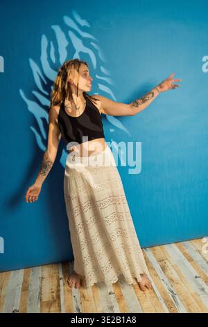 Jeune femme avec des tatouages et des dreadlocks posant dans une longue jupe sur un fond bleu Banque D'Images