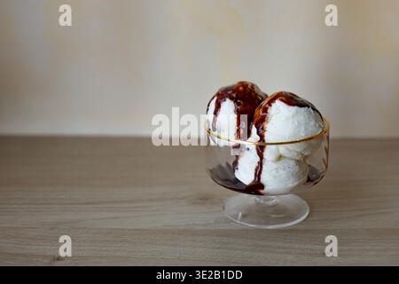 des boules de crème glacée aux haricots de vanille servies dans un bol à dessert en verre transparent avec un bord doré. Le dessert est arrosé de sirop de chocolat noir riche ou de fudge Banque D'Images
