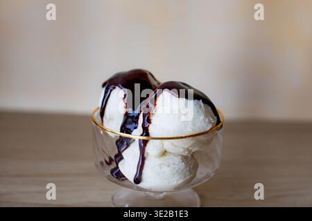 des boules de crème glacée aux haricots de vanille servies dans un bol à dessert en verre transparent avec un bord doré. Le dessert est arrosé de sirop de chocolat noir riche ou de fudge Banque D'Images
