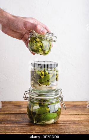 Nourriture, main masculine tenant l'un des trois pots en verre de poivrons jalapeno fraîchement marinés avec des herbes et de l'ail. Espace de copie Banque D'Images