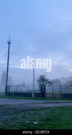 Deux tours de communication se tiennent hautes dans le brouillard à côté d'un terrain de sport L'environnement est tôt le matin avec une faible visibilité et une atmosphère calme surro Banque D'Images