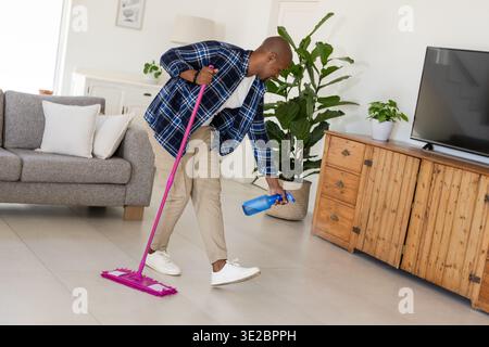 Homme afro-américain nettoyant le salon, se penchant vers l'avant tenant une serpillière rose et un vaporisateur bleu. Maison, adulte, mâle, rangé, plancher, tuiles, plantes Banque D'Images