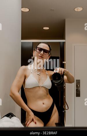 Femme se tient devant un miroir prenant une photo tout en portant un bikini et des lunettes dans une salle de bain avec des couleurs neutres et un éclairage doux Banque D'Images