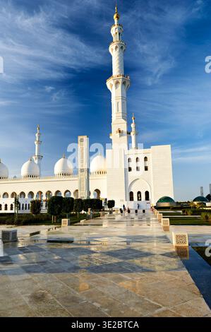 Les gens se promènent autour de la Grande Mosquée Sheikh Zayed à Abu Dhabi. Le soleil se couche derrière la mosquée créant des couleurs chaudes dans le ciel. Banque D'Images