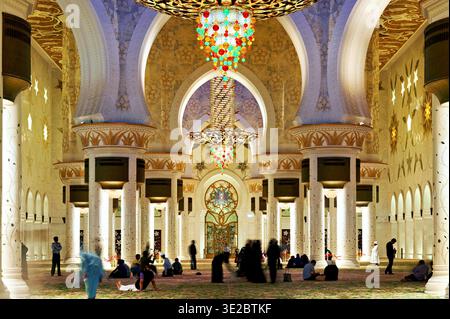 Les gens marchent et sont assis dans la Grande Mosquée Sheikh Zayed à Abu Dhabi. L'espace est bien éclairé avec des décorations ornées uniques. Banque D'Images