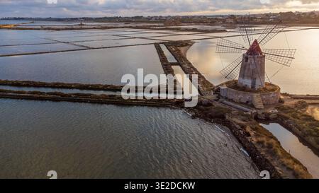 Une vue aérienne époustouflante capture le charme intemporel des marais salants de Marsala. Un ancien moulin à vent se dresse parmi les bassins géométriques, tandis que le coucher de soleil doré se lève Banque D'Images