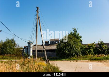 Des poteaux électriques en bois penchés et une boîte de transformateur Janky avec un désordre de fils se tiennent près d'une vieille route de campagne. Bâtiments rustiques au loin sous le bleu Banque D'Images