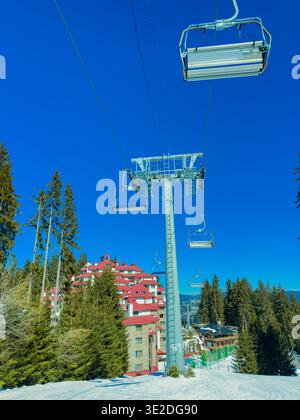 Une station de ski se trouve en arrière-plan avec un télésiège s'élevant au-dessus d'un sol enneigé et de grands arbres sous un ciel bleu clair. Les gens aiment le ski et le Banque D'Images
