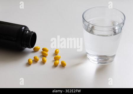 Pilules jaunes renversées d'une bouteille sombre à côté d'un verre d'eau sur un fond blanc. Soins de santé, vitamines, médicaments et concept de supplément quotidien Banque D'Images