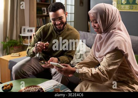 Jeune homme musulman adulte et jeune femme musulmane adulte assis sur un canapé partageant le thé et regardant un smartphone ensemble, souriant et s'engageant dans une conversation joyeuse Banque D'Images