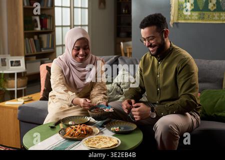 Jeune femme musulmane adulte portant le hijab et jeune homme musulman adulte assis sur un canapé partageant ensemble un repas traditionnel, souriant et interagissant avec la nourriture Banque D'Images