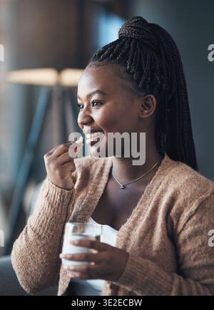 Femme noire heureuse et pilule avec de l'eau à la maison pour des suppléments, le système immunitaire ou des médicaments. Verre liquide, personne et sourire avec capsule dans la maison pour Banque D'Images