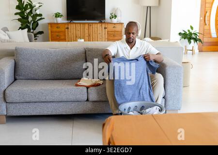 Homme afro-américain pliant le linge tout en tenant le tee-shirt bleu au-dessus du panier rond sur le canapé, espace de copie Banque D'Images