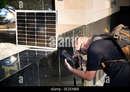 Un photographe masculin prend des photos détaillées, avec un panneau solaire installé à proximité. Sac à dos indique la mobilité, mettant en évidence l'utilisation des énergies renouvelables dans la photographie. Énergie solaire pour une charge écologique. Banque D'Images