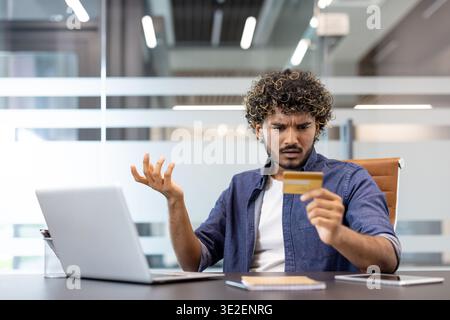 Un jeune Indien est assis à un bureau dans un bureau et regarde avec déception la carte de crédit qu'il tient dans sa main. Banque D'Images