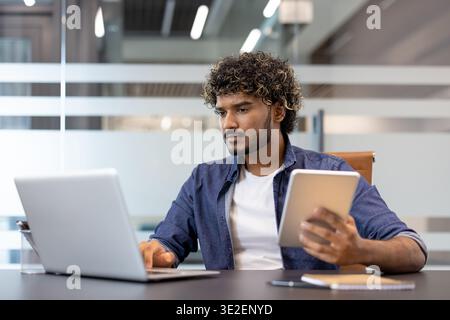Un jeune homme d'affaires indien concentré est assis à son bureau dans le bureau, tenant une tablette à la main et travaillant sur un ordinateur portable. Banque D'Images
