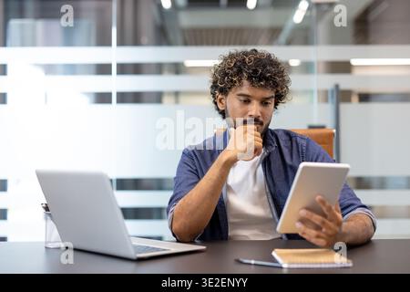 Portrait d'affaires d'un jeune homme indien travaillant sérieusement et soigneusement au bureau à l'aide d'une tablette. Banque D'Images