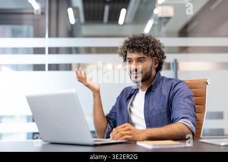 Un jeune indien est assis dans un bureau devant un ordinateur portable et parle lors d'un appel vidéo. Banque D'Images