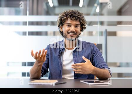 Portrait d'un bel homme indien assis dans un bureau à un bureau devant la caméra, parlant et faisant des gestes avec ses mains. Banque D'Images