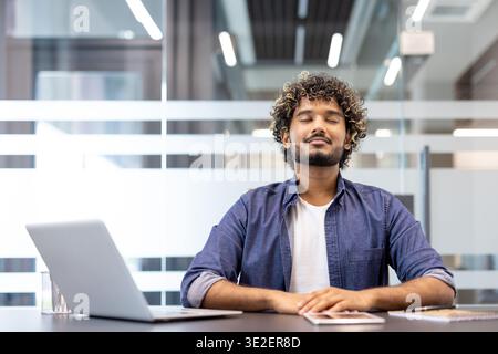 Un jeune homme indien calme et concentré est assis à un bureau dans le bureau, les yeux fermés, se reposant avec soin. Banque D'Images
