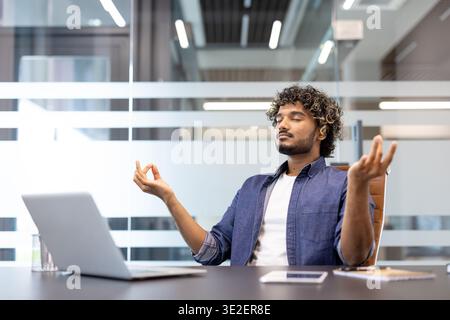 Détendu et calme jeune homme indien assis dans une chaise de bureau à un bureau dans une position de lotus et méditant. Banque D'Images
