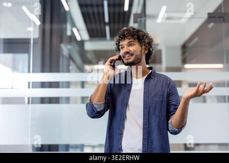 Homme indien souriant debout dans le bureau et parlant émotionnellement au téléphone, faisant des gestes avec ses mains. Banque D'Images