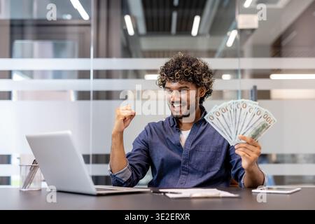 Heureux jeune homme indien assis dans le bureau, regardant l'écran de l'ordinateur portable, tenant de l'argent liquide dans ses mains. Banque D'Images