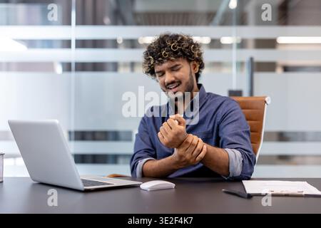 Jeune homme indien assis à son lieu de travail dans le bureau et tenant son poignet avec sa main, ressentant de la douleur. Banque D'Images