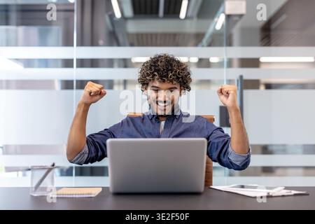 Heureux jeune homme indien assis à son lieu de travail dans le bureau, se réjouissant du succès, regardant l'écran de l'ordinateur portable et levant les mains, célébrant. Banque D'Images